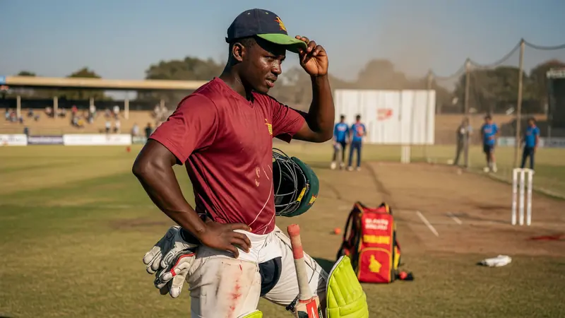 Cricketer in a maroon training shirt and dark cap adjusts his cap while holding a bat and helmet on a sunlit practice ground