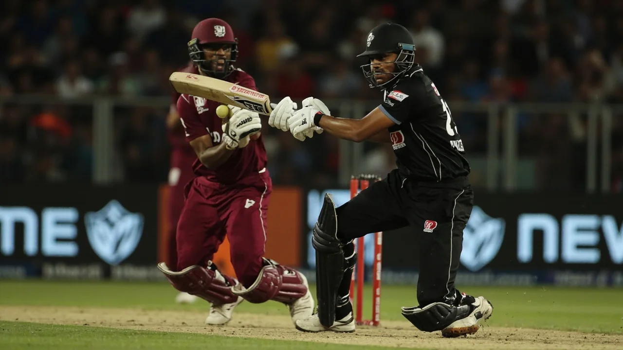 Tight sideline action frame of a batter playing a horizontal-bat shot at the crease with the ball visible just after contact, a slip fielder in the foreground and the Providence stands rising behind the rope